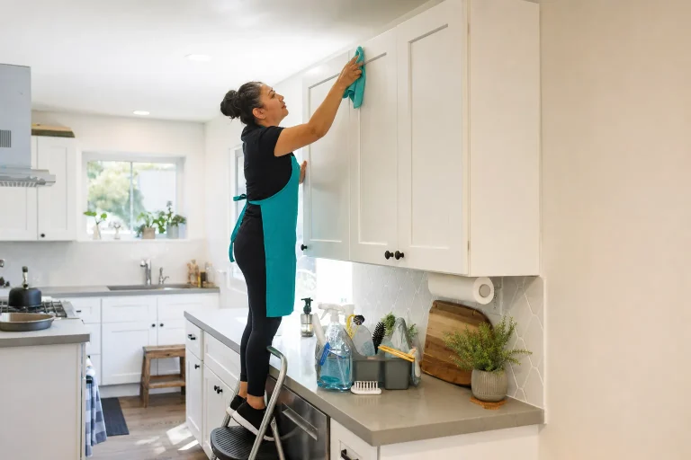 Cleaner in a teal apron deep cleaning the tops of kitchen cabinets in a Gresham home